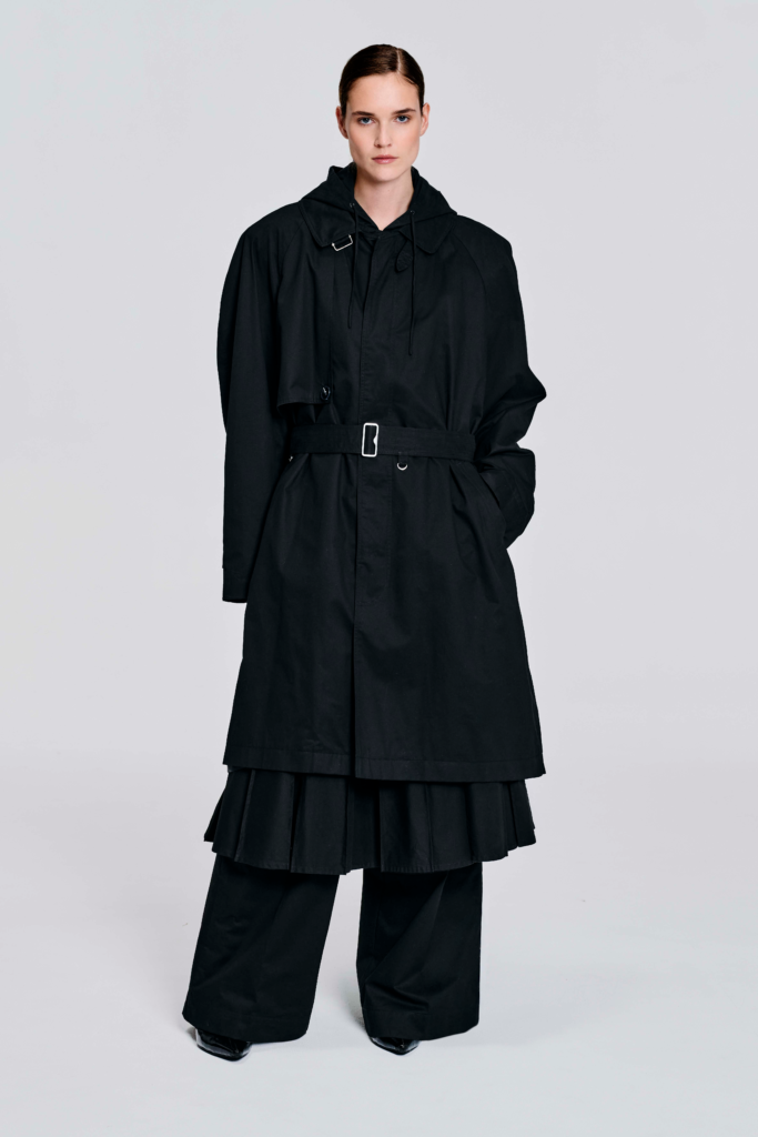 Model wearing a black hooded cotton coat with oversized longline silhouette, drawstring hood, wide sleeves, cuff tabs, and back strap detailing by KimC Studio.