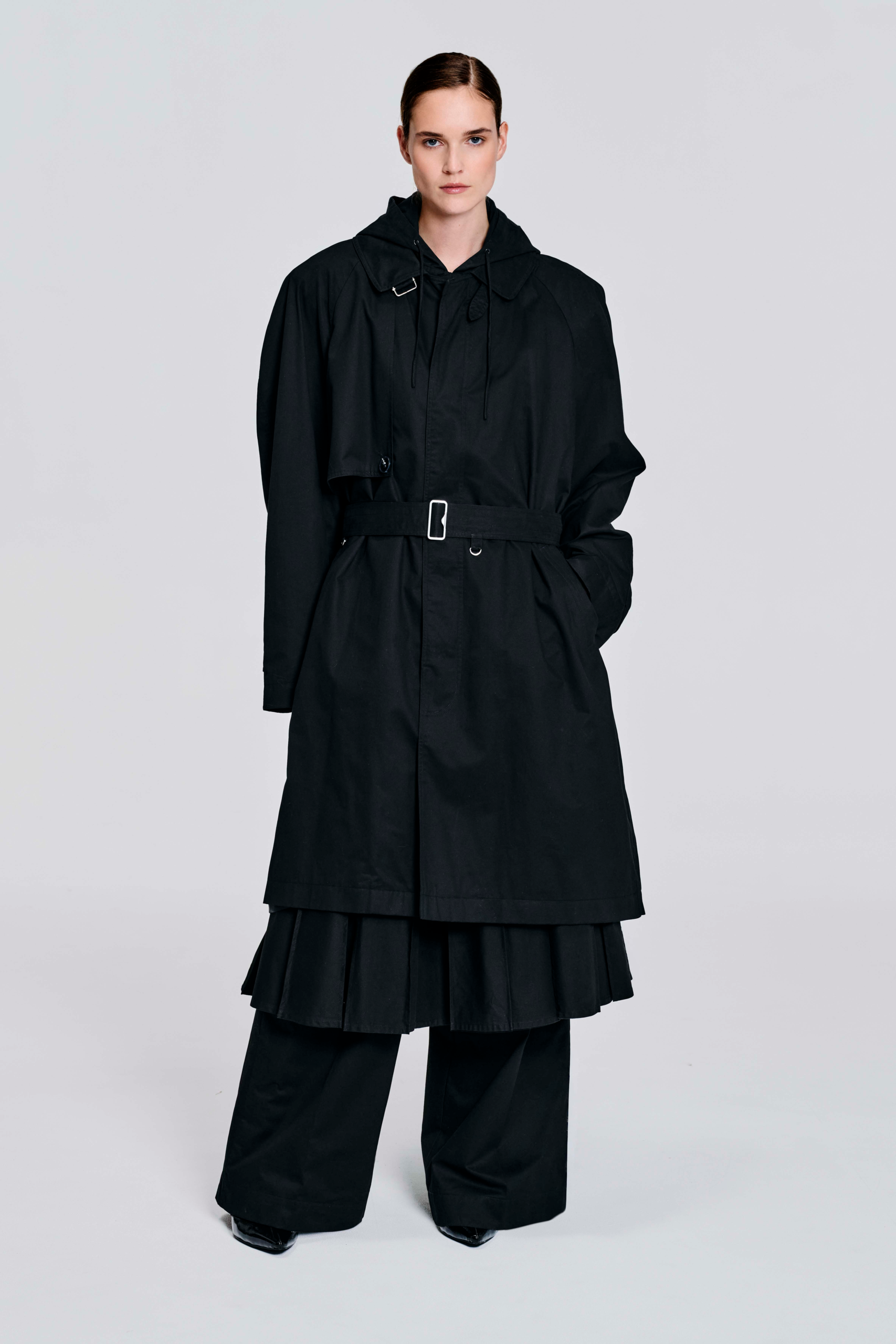 Model wearing a black hooded cotton coat with oversized longline silhouette, drawstring hood, wide sleeves, cuff tabs, and back strap detailing by KimC Studio.