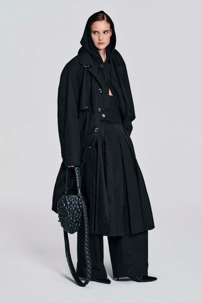Model wearing a black hooded cotton coat with oversized longline silhouette, drawstring hood, wide sleeves, cuff tabs, and back strap detailing by KimC Studio.